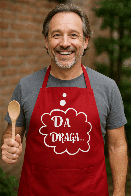 Crno-bijela fotografija sretno muškarca u kuhinji u kućnoj odjeći s natpisom 'Da Draga' na crvenoj pregači, drži drvenu žlicu, uz prirodno zeleno okruženje.